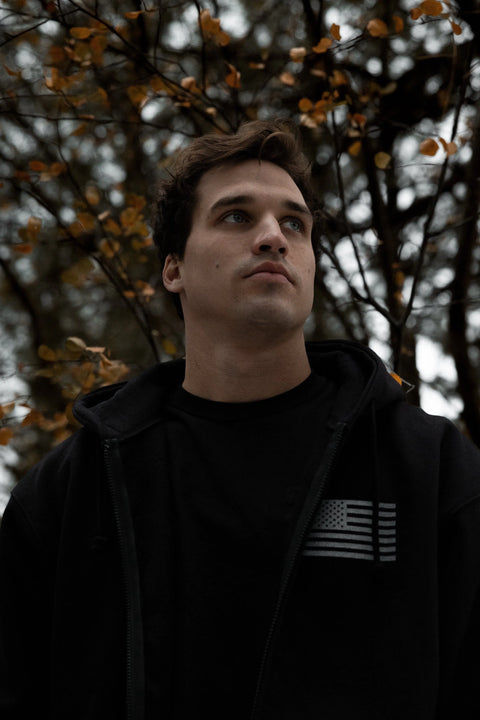 A young man in a Proudly USA American Flag - Zipper Hoodie Sweatshirt (Black) gazes upward. Blurred autumn leaves and branches form the background, with soft lighting emphasizing its premium, made-in-USA details.