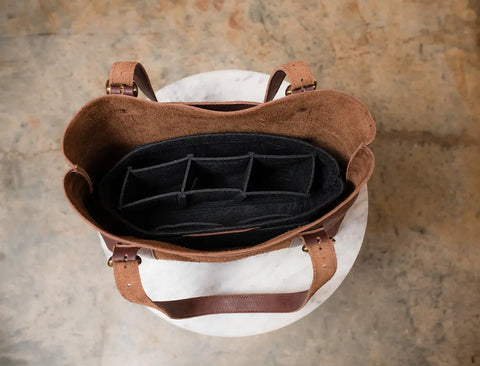 A top-down view of an open brown leather handbag with the Urban Southern Tote Insert, a black lightweight felt organizer, inside. The bag is set on a round white marble surface with a brown textured background.
