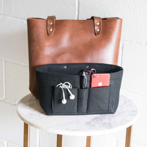 An Urban Southern Tote Insert sits in front of a brown leather tote bag on a round marble table, offering customizable organization for earphones, phone, sunglasses, and pens, with a white brick wall in the background.