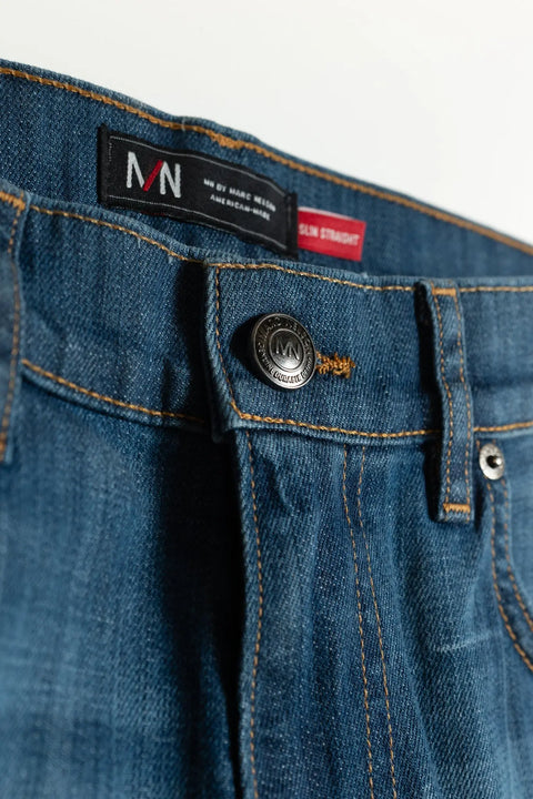 Close-up of The Nelson Medium Wash Denim jeans by Marc Nelson, featuring blue denim, a metal M-engraved button, orange stitching, and a black M|N by Marc Nelson label inside the waistband. Made in the USA.