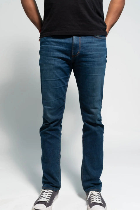 A person, visible from the waist down, stands against a plain background with hands in pockets, wearing Marc Nelsons The Nelson Medium Wash Denim jeans in blue, paired with a black t-shirt and gray sneakers.