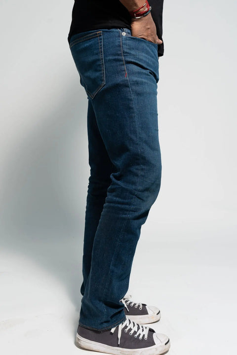 A person stands sideways against a plain white background, one hand in their pocket, wearing Marc Nelsons The Nelson Medium Wash Denim jeans, paired with a black shirt and black-and-white sneakers.