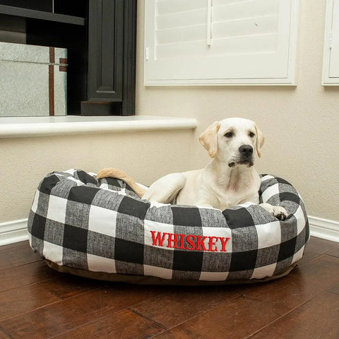 A yellow Labrador rests in a water-resistant, black-and-white Anderson Check Bagel Bed by Majestic Pet, personalized with WHISKEY in red, placed on a wooden floor near a window with white shutters.