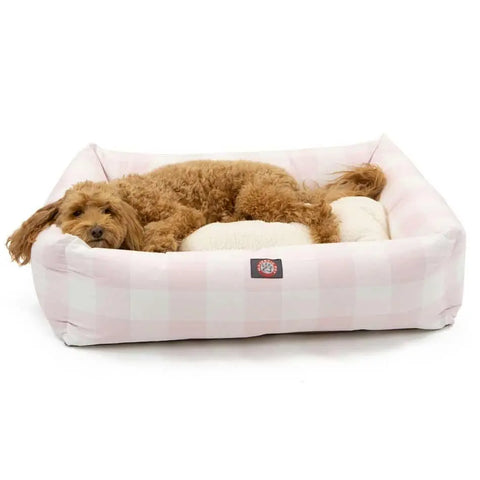 A small, fluffy brown dog is lying comfortably in the Majestic Pet Anderson Check Luxe Bagel Bed, featuring a light pink and white checkered design and a cream-colored faux sheepskin cushion.