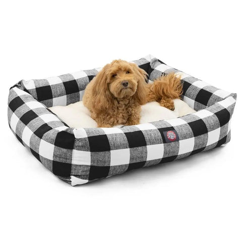 A small, fluffy brown dog is resting in the Majestic Pet Anderson Check Luxe Bagel Bed, which features a soft cream faux sheepskin cushion and a black-and-white plaid rectangular frame with a machine washable slipcover.