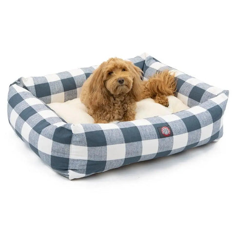 A small, fluffy brown dog relaxes in the Majestic Pet Anderson Check Luxe Bagel Bed, featuring a blue and white checkered machine washable slipcover and a soft cream faux sheepskin cushion.