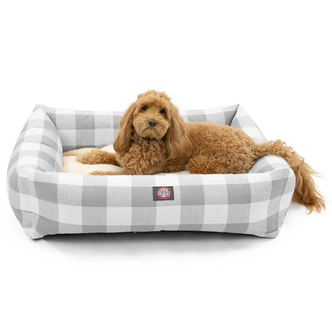 A curly-haired brown dog is lying comfortably in the Majestic Pet Anderson Check Luxe Bagel Bed, featuring soft sides, a gray and white checkered machine-washable slipcover, and a logo on the front.