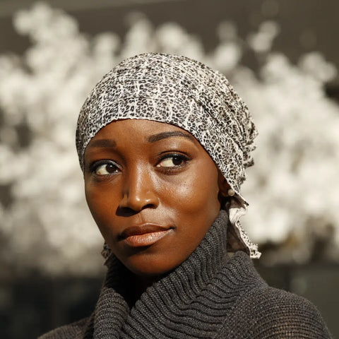A woman in a Gina Chemo Beanie® by Chemo Beanies® and a dark sweater gazes thoughtfully to the side, with white blossoms softly blurred in the background.