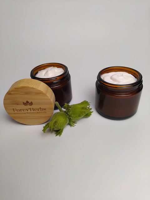 Two brown glass jars of Forev'Herbs Anti-Aging Facial Cream sit on a white surface. A bamboo lid with the Forev'Herbs logo rests nearby, while fresh green hazelnuts and Tremella add a natural accent between the jars and lid.