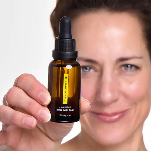 A light-skinned, brown-haired person is smiling and holding up a brown glass dropper bottle labeled Essentials Superset by Open Formula, an anti-aging skincare essential, in front of their face against a white background.