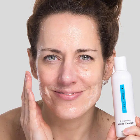 A woman with wet, glowing skin smiles as she applies Open Formula’s Transformational Routine cleanser—an essential part of her anti-aging skincare regimen—while holding the sleek white bottle against a simple light backdrop.