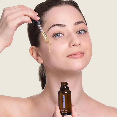 A woman applies Open Formulas Transformational Routine serum to her cheek with a dropper, holding the brown bottle in her other hand. She has clear skin, light eyes, and brown hair pulled back, smiling slightly at the camera.