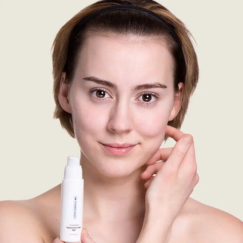 A woman with short brown hair and a black headband smiles softly as she touches her face and holds a white Open Formula Transformational Routine bottle against a light beige background.