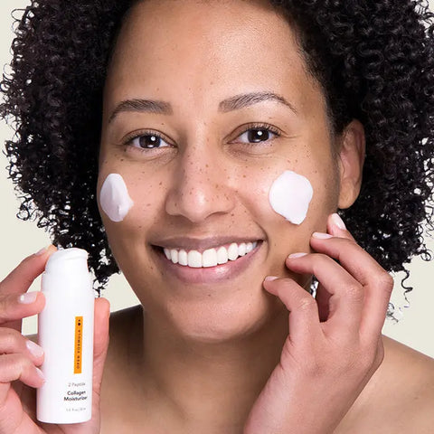 A smiling woman with curly hair, clear skin, and freckles applies Open Formula’s Transformational Routine collagen and anti-aging moisturizer from a white bottle to her cheeks.