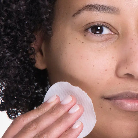 A person with curly hair is gently pressing a round cotton pad to their cheek while using Open Formulas Transformational Routine, suggesting an effective anti-aging skincare or makeup removal step. Only the lower half of the face and part of the eye are shown.