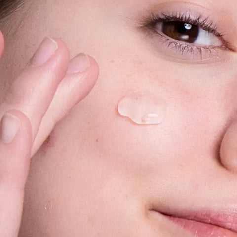 A close-up of a person applying Open Formula Transformational Routine Vitamin C serum to their cheek with their fingers, showing part of their eye, nose, and lips for an anti-aging skincare routine.