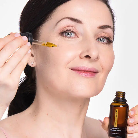 A woman with fair skin and dark hair smiles as she applies Open Formulas Transformational Routine serum to her cheek, holding the dropper near her face and the bottle in her other hand.