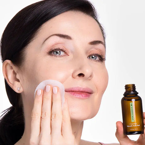 A woman with fair skin is smiling and applying skincare to her cheek with a cotton pad. She holds a brown bottle labeled Open Formula Anti-Aging Superset in her other hand.