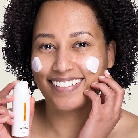 A woman with curly hair smiles at the camera, applying Open Formulas Anti-Aging Superset to her cheeks. She holds a white skincare bottle in one hand and has two dabs of the product on her face.
