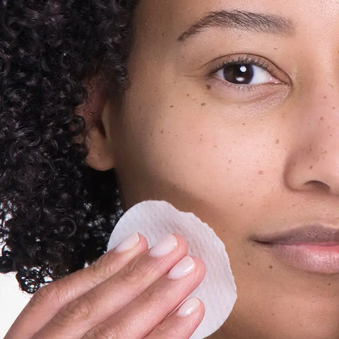 A person with curly hair gently presses a cotton pad to their cheek, showing skincare or makeup removal—possibly after using Open Formula’s Anti-Aging Superset. Only the lower half of their face is visible in close-up.