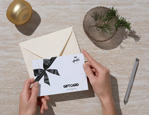 A person holding a Apollo gift card with a black ribbon bow on it, with a envelope, pen, and decorative items in the background.
