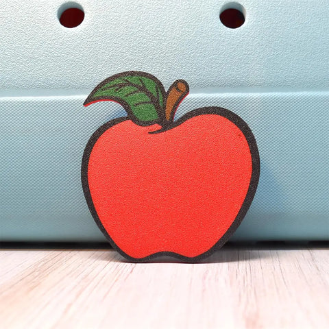 The Chatelet Manufacturing FRESHe Baglets Apple Teacher Charm is a flat, red apple with a green leaf and brown stem on a light blue, textured background—perfect as a decorative tote bag accessory or thoughtful teacher gift. Compatible with popular tote bags.