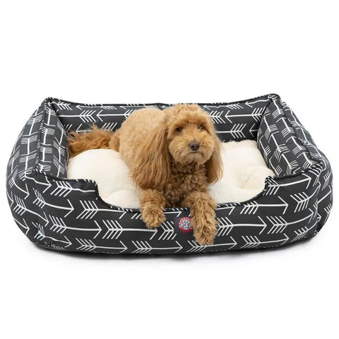 A small, curly-haired brown dog relaxes on a plush cream cushion inside the Majestic Pet Arrow Contemporary Lounge Bed—a black rectangular bed with white arrow patterns that blends modern style with cozy comfort.