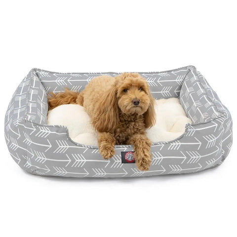 A small, curly-haired brown dog relaxes on the Majestic Pet Arrow Contemporary Lounge Bed, featuring a plush cream surface and a gray cover with white arrow designs. The dog is lying down and facing forward.