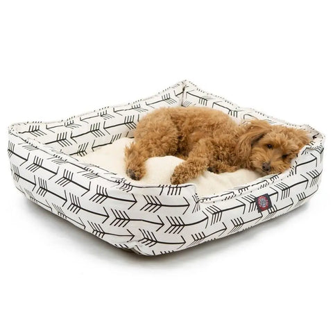 A small, curly-haired brown dog rests comfortably on the plush surface of the Majestic Pet Arrow Contemporary Lounge Bed, featuring a stylish arrow pattern in black and white.