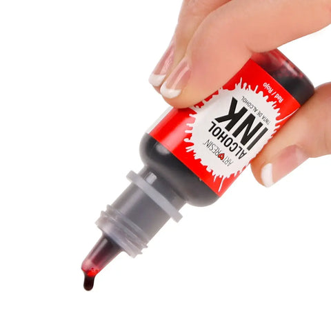 A hand holds a red bottle of ArtResin Alcohol Ink by ArtResin, with a drop poised at the nozzle. The label displays a splash design and offers vibrant color, ideal for resin art and other creative projects.