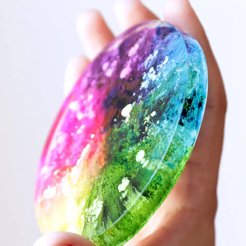A hand holds a round, clear resin disc made with ArtResin Alcohol Ink by ArtResin, featuring vibrant pink, purple, green, and blue shades blending together and white cloudy patterns inside.