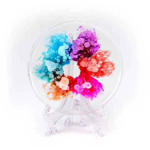 A round glass plate on a clear stand, featuring vibrant blue, purple, pink, orange, and red hues in a splattered circular pattern. Inspired by resin artists and the dynamic effects created with ArtResin Alcohol Ink by ArtResin.
