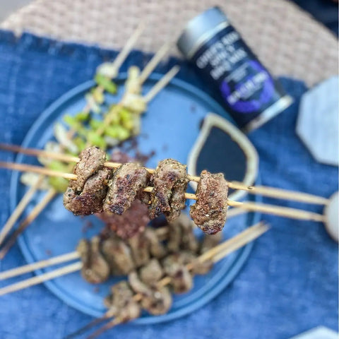 Close-up of grilled meat skewers with veggies on a blue plate, paired with a spice container from the Gustus Vitae Bougie BBQ Essentials Collection - 3 Pack, all arranged on a blue textured cloth.