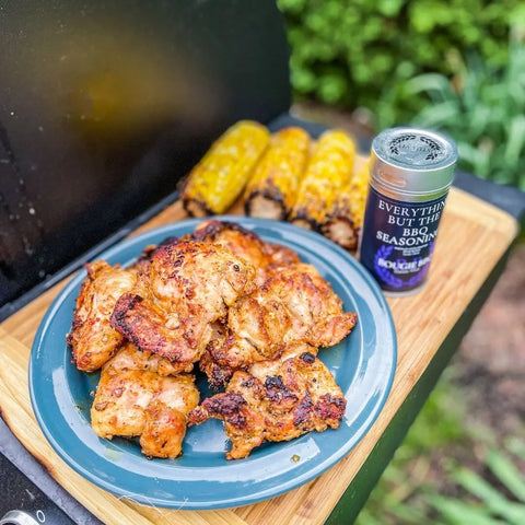A blue plate of grilled chicken thighs sits next to corn and a Gustus Vitae Bougie BBQ Essentials Collection - 3 Pack, all by an open grill outdoors with greenery in the background.