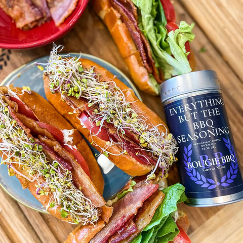 Three sandwiches with bacon, lettuce, tomato, and sprouts sit on a wooden surface beside a tin from the Gustus Vitae Bougie BBQ Essentials Collection - 3 Pack. A red plate with sandwich halves is also in view.