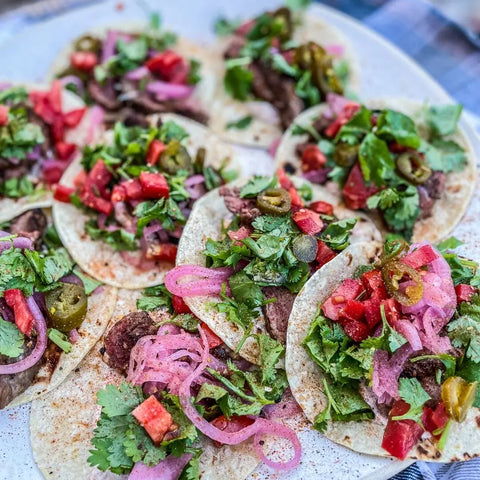 A plate of vibrant small tacos topped with chopped beef, cilantro, tomatoes, pickled red onions, and jalapeños is finished with a sprinkle from the Gustus Vitae Gourmet Grilling Salts Collection - 3 Tins for extra flavor.