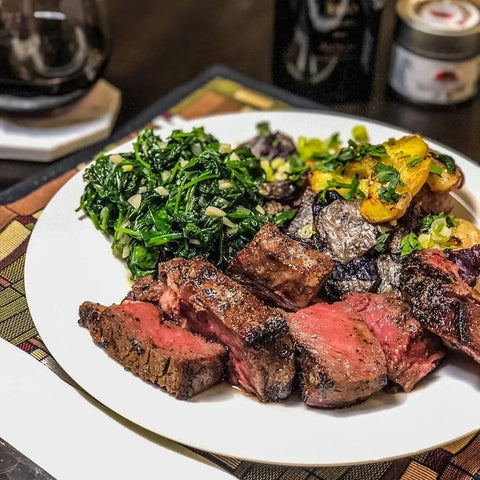 A plate of grilled steak slices seasoned with Gustus Vitaes Gourmet Grilling Salts Collection, served alongside sautéed greens and roasted multicolored potatoes, sits on a patterned placemat, with red wine and condiments in the background.