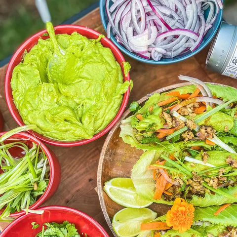 A vibrant spread with guacamole, sliced red onions, herbs, veggie lettuce wraps, lime wedges, and an edible orange flower—finished with Gustus Vitae Gourmet Grilling Salts Collection - 3 Tins on a wooden surface.