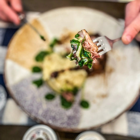 A close-up of a fork with food seasoned using the Gustus Vitae European Seasonings Gift Set - Tastes of Europe, above mashed potatoes and sauce. Blurred hands and plate in the background, on a blue and white tablecloth.