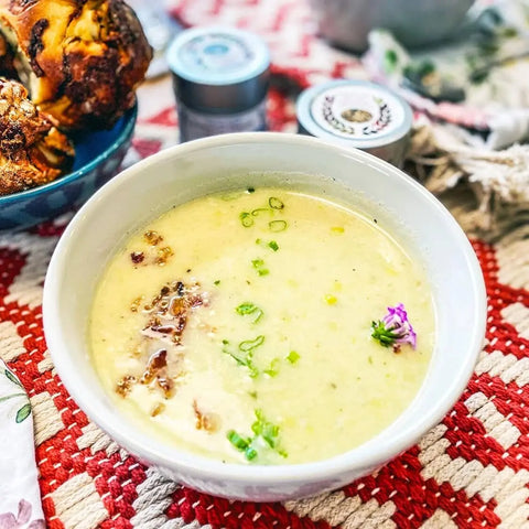A bowl of creamy soup garnished with green onions and a purple flower sits on a red and white woven cloth, with roasted meat and the Gustus Vitae European Seasonings Gift Set - Tastes of Europe artisanal spice blends six pack in the background.