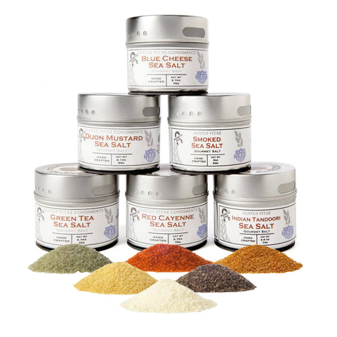 The Gustus Vitae Painters Palette Collection features six tins of different finishing sea salts stacked in a pyramid, with vibrant piles of each colorful salt displayed in front.