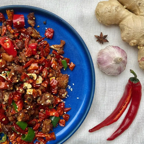 A blue plate of spicy stir-fried meat sits near fresh ginger, garlic, and chili—perfectly enhanced by Gustus Vitae’s Painter’s Palette Collection - 6 Tins gourmet sea salts for an extra boost of flavor.