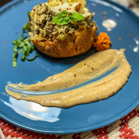 A gourmet baked stuffed potato topped with herbs and gourmet sea salts from Gustus Vitae’s Painters Palette Collection - 6 Tins, served on a blue plate with fresh basil, shredded greens, a brown sauce smear, and an orange garnish.