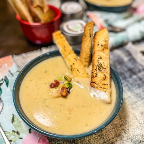 A bowl of creamy soup topped with green onions and crispy bacon is served with three toasted breadsticks. In the background: breadsticks and the Gustus Vitae French Seasoning Gift Set, featuring six artisanal Tastes of France spice blends.