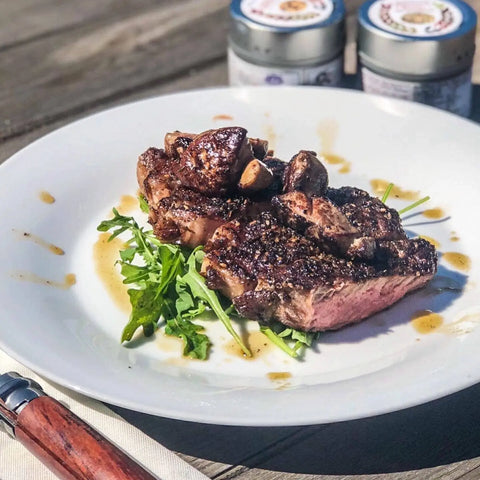 A seared steak rests on arugula, drizzled with sauce and Gustus Vitae’s French Seasoning Gift Set - Tastes of France artisanal spices. Two tins from the six-pack and a knife are in the background on a wooden table.