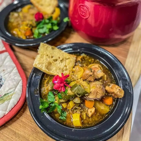A hearty stew featuring meat, vegetables, and herbs seasoned with Gustus Vitae’s French Seasoning Gift Set – Tastes of France artisanal spice blends, garnished with parsley and a pink flower, served with bread on a wooden surface.