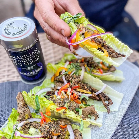 A hand picks up a lettuce wrap filled with seasoned beef, red onions, and bell peppers. Three more wraps sit on a plate next to a container of Gustus Vitae Global Gourmet BBQ Collection - 4 Pack seasoning.