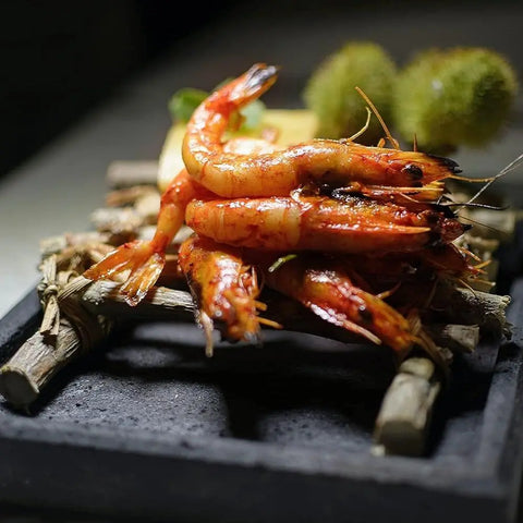 Grilled shrimp glazed with Gustus Vitaes Global Gourmet BBQ Collection - 4 Pack, stacked on a wooden tray, garnished with herbs, and served on a black stone slab in an artistic presentation.