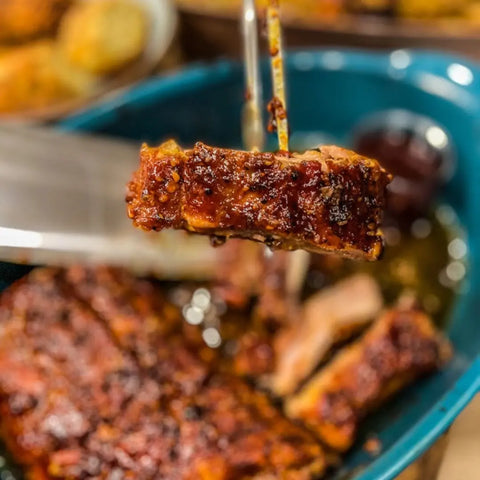 A close-up of succulent ribs with a caramelized crust, held above a pan of more ribs, highlights flavors from the Gustus Vitae Global Gourmet BBQ Collection - 4 Pack artisanal rubs.