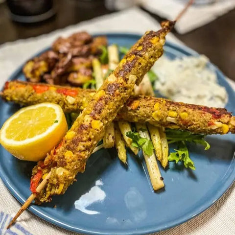 Two grilled vegetable kebabs, seasoned with Gustus Vitaes Greek Seasoning Gift Set - Tastes of Greece, served on asparagus and greens with sliced lemon, sautéed mushrooms, and creamy white sauce on a blue plate.